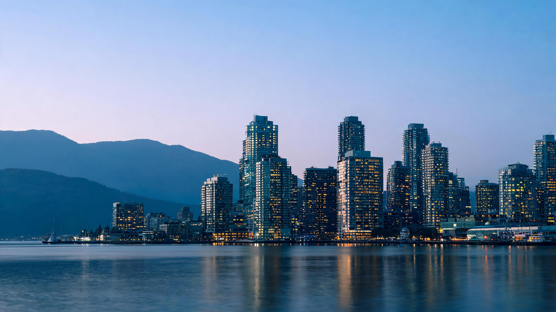 A Canadian city skyline at dusk seen across the harbour, towers lit from within with coastal mountains behind