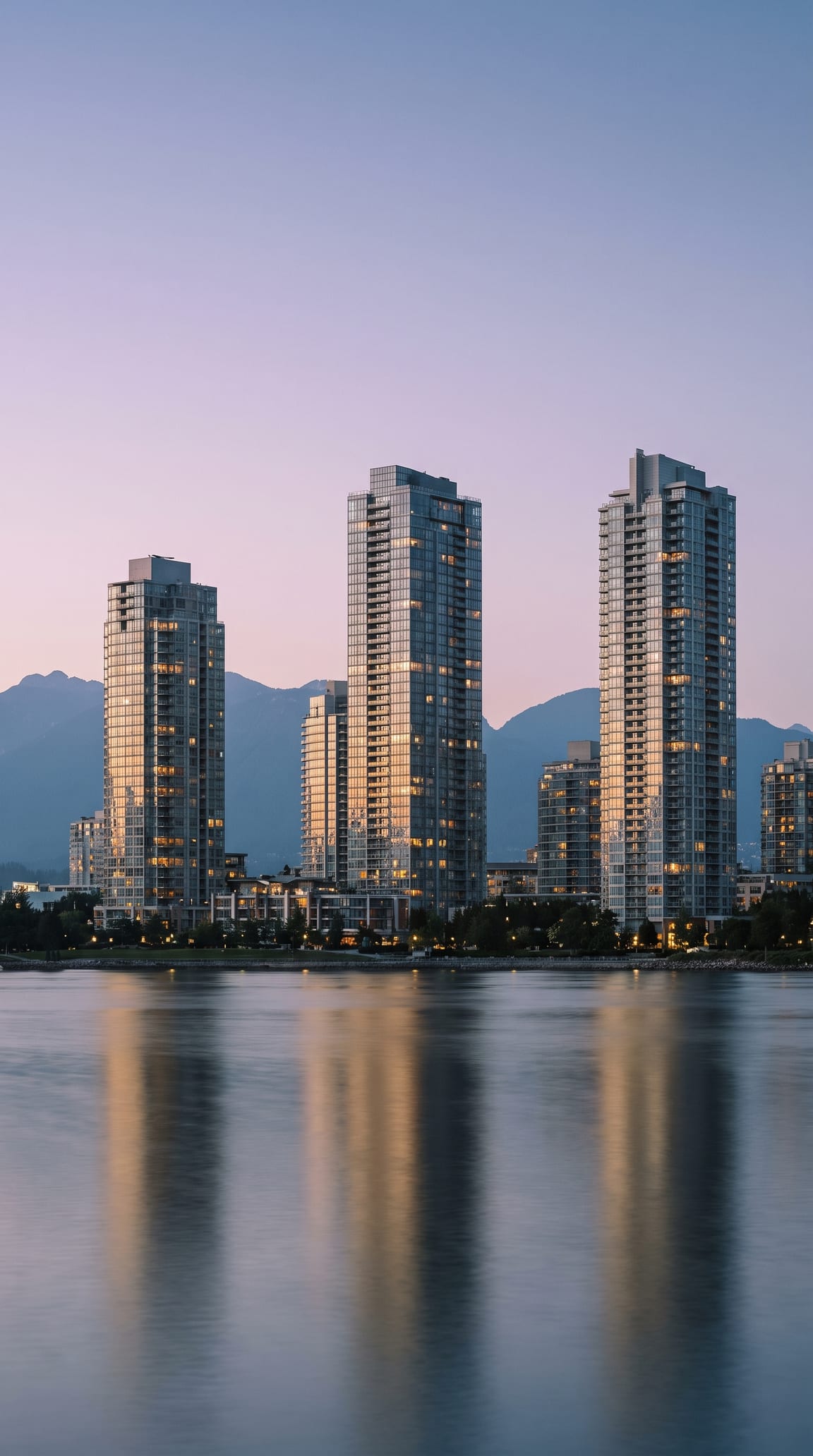 A Canadian waterfront city at blue hour, glass towers lit from within above still harbour water, coastal mountains behind